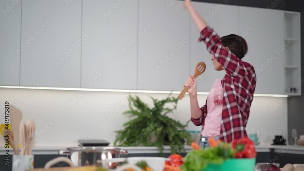 Woman dancing and singing using wooden spoon preparing food at new home kitchen, carefree joyful housewife having fun cooking dinner meal listen to music in modern kitchen. FHD footage. Woman dancing and singing using wooden spoon preparing food at new home kitchen, carefree joyful housewife having fun cooking dinner meal listen to music in modern kitchen. FHD footage.