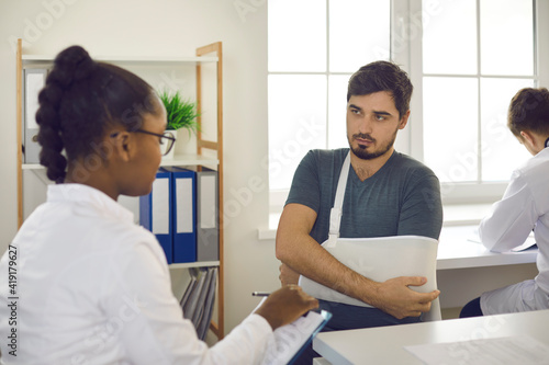 Caucasian man with hand in bandage sling pointing to injured elbow complains of arm pain at doctor appointment. African american woman specialist listening carefully to patient with hand trauma