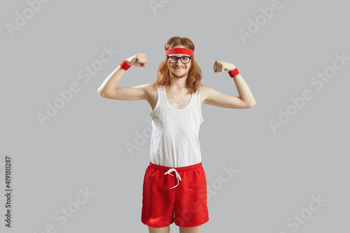 Funny thin skinny man with long hair in eyeglasses, retro headband, white tank top and red shorts showing weak muscles standing isolated on gray background. Sports workout and fitness exercise concept