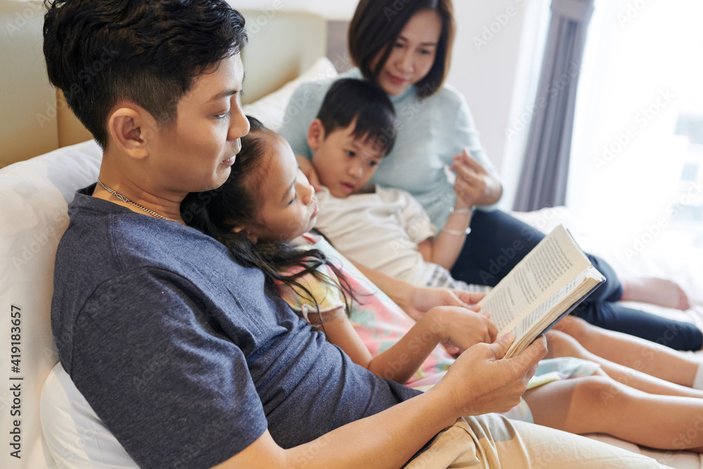 Vietnamese man reading interesting book for his children and wife whe they all are lying on bed Vietnamese man reading interesting book for his children and wife whe they all are lying on bed