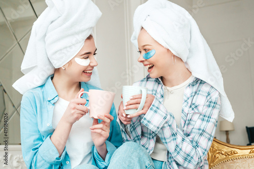 Two young beautiful smiling women with patches under their eyes.Sexy carefree models sitting on bed in posh apartment.They doing beauty treatments at home in towels on heads. Drinking tea  holding cup