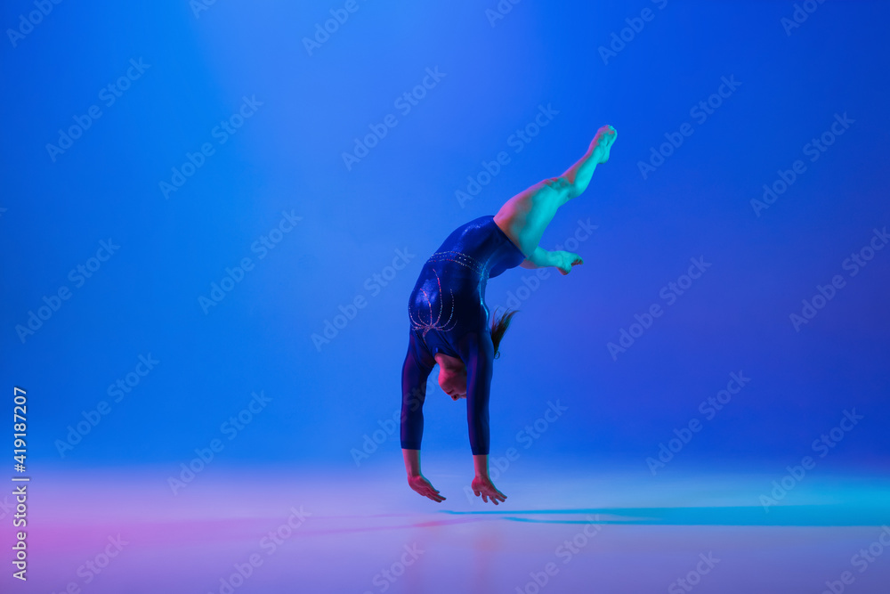 Energy. Young flexible girl isolated on blue studio background in neon light. Young female model practicing artistic gymnastics. Exercises for flexibility, balance. Grace in motion, sport, action.