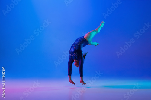 Energy. Young flexible girl isolated on blue studio background in neon light. Young female model practicing artistic gymnastics. Exercises for flexibility, balance. Grace in motion, sport, action.