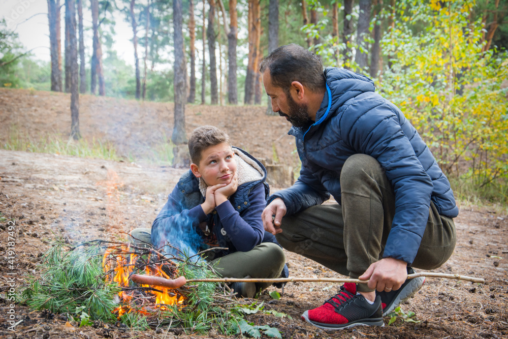 In the fall, father and son sit in the forest near the fire and fry sausages. In the fall, father and son sit in the forest near the fire and fry sausages.