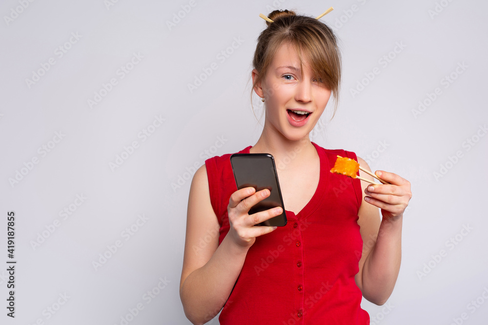 Blond-haired girl eating sushi with chopsticks, isolated on gray background with side space. Young woman holding smartphone ready to place another order, online delivery Blond-haired girl eating sushi with chopsticks, isolated on gray background with side space. Young woman holding smartphone ready to place another order, online delivery