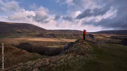 mountaineer in red jacket on a hill on the isle of skye, scotland looking towards a waterfall in a mountainous landscape. cloudy sky