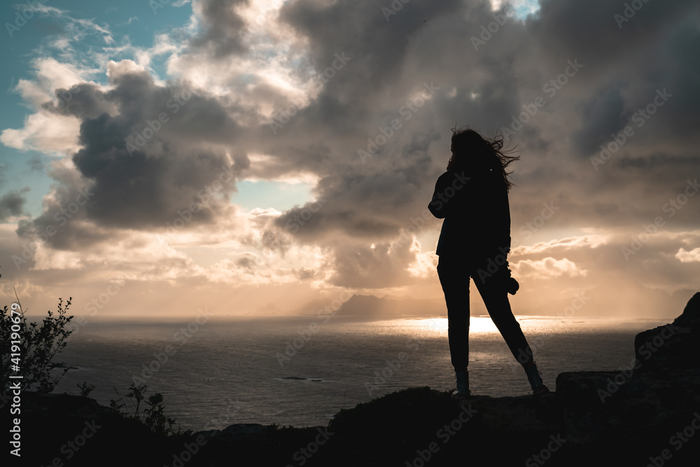 A woman stands with wind blown hair silhouette at the top of a mountain during sunset. Excellent adventure and woman accomplishment shot