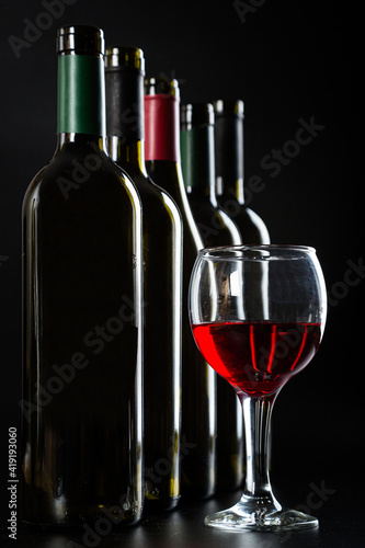 several different bottles of wine and a glass with red wine on a dark glossy background.