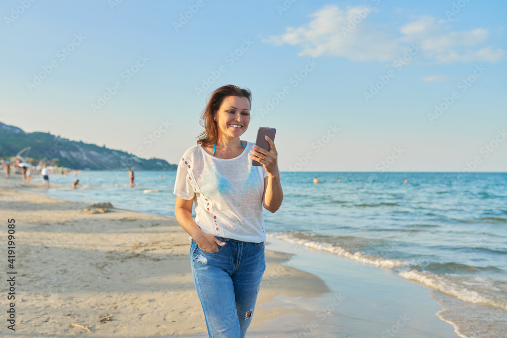 Middle-aged woman walking along beach with smartphone Middle-aged woman walking along beach with smartphone