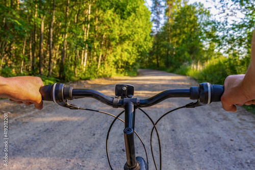 Human hands holding bicycle handlebar against landscape with country road. Man travelling by bicycle in northern forest in summer evening. Weekend activities, healthy lifestyle, adventure concept