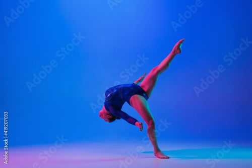 Energy. Young flexible girl isolated on blue studio background in neon light. Young female model practicing artistic gymnastics. Exercises for flexibility, balance. Grace in motion, sport, action.