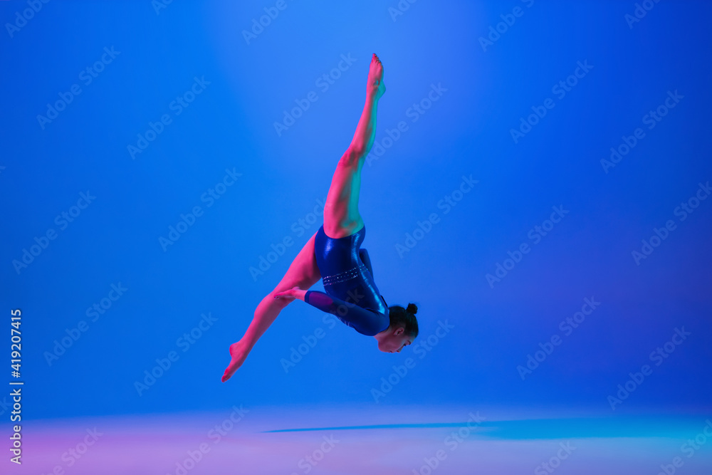 Flying high. Young flexible girl isolated on blue studio background in neon light. Young female model practicing artistic gymnastics. Exercises for flexibility, balance. Grace in motion, sport, action