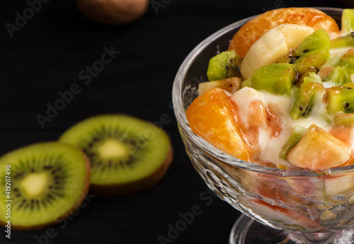 tropical fruit salad in glass bowl. Kiwi, banana and tangerine with yogurt and topping. Veggie dessert. Healthy food concept. Black wood background. High quality photo
