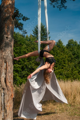 A slender girl with blonde hair, in a black stage costume, performs gymnastic and circus exercises on white silk against the background of the forest and the sky.