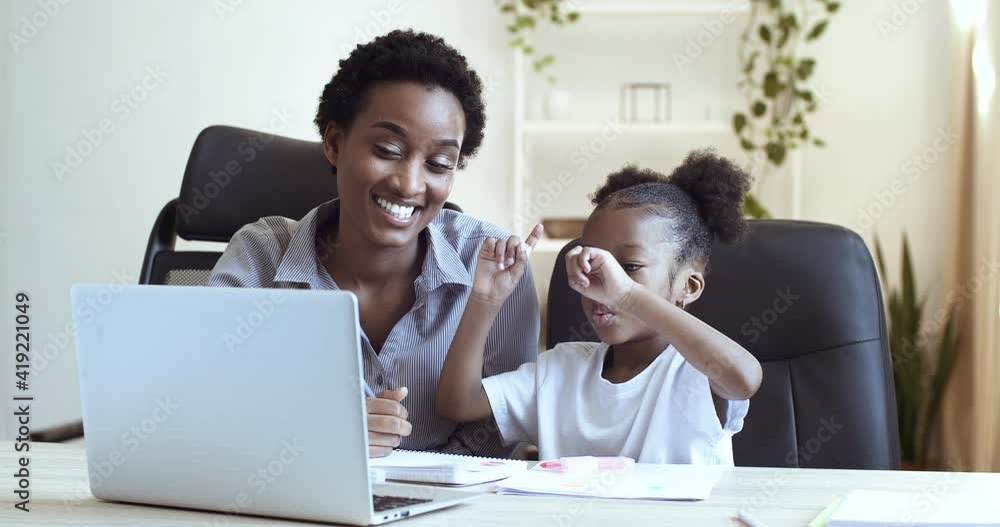 Young mother african american woman teacher with cute daughter schoolgirl black child sitting at table in front of laptop study remotely doing homework online in network laughing smile close-up Young mother african american woman teacher with cute daughter schoolgirl black child sitting at table in front of laptop study remotely doing homework online in network laughing smile close-up