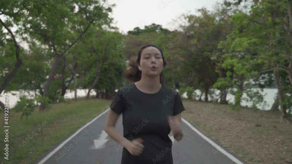 asian athlete woman in black sportswear and wireless headphone running in urban city trees park during sunset and bad cloudy weather. female jogging outdoor in evening. healthy workout. front view. asian athlete woman in black sportswear and wireless headphone running in urban city trees park during sunset and bad cloudy weather. female jogging outdoor in evening. healthy workout. front view.