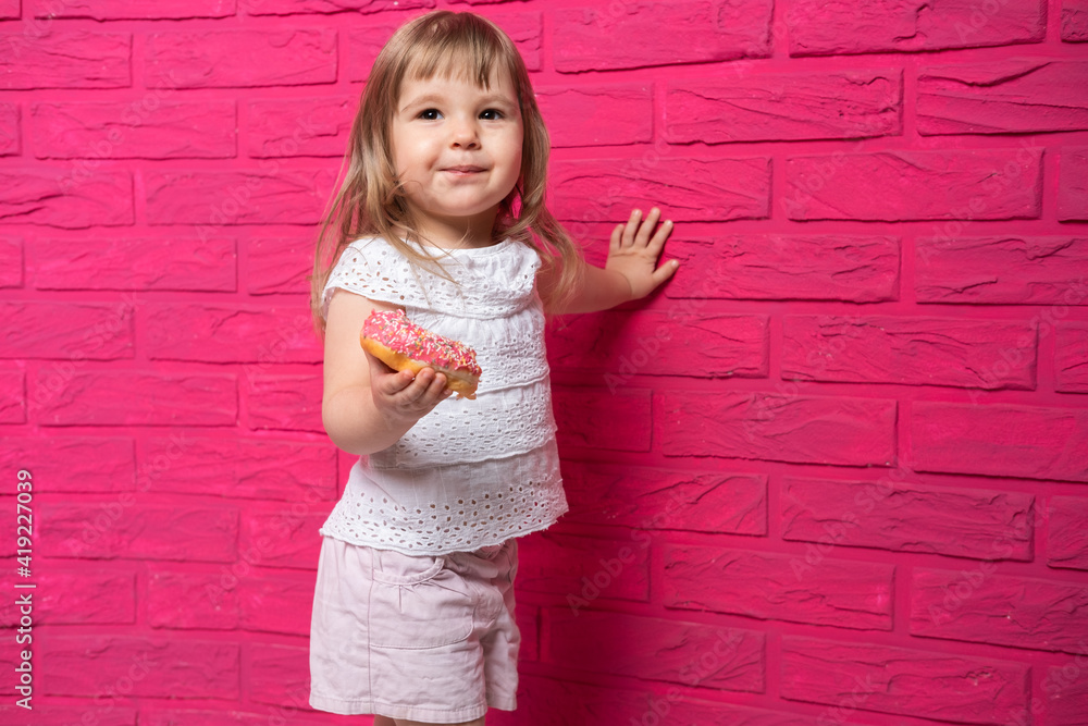 little funny blonde girl in white shirt eats donut on pink background. little funny blonde girl in white shirt eats donut on pink background.