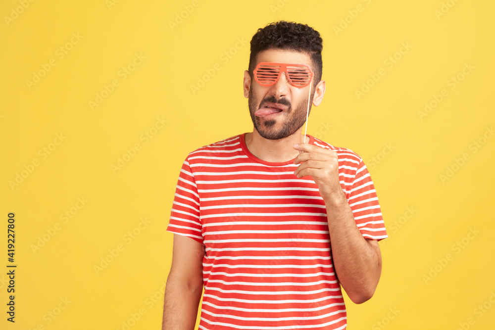 Funny positive man with beard in striped t-shirt fooling around and having fun, showing tongue holding red eyeglasses mask on stick near eyes. Indoor studio shot isolated on yellow background Funny positive man with beard in striped t-shirt fooling around and having fun, showing tongue holding red eyeglasses mask on stick near eyes. Indoor studio shot isolated on yellow background