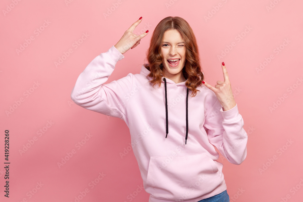 Portrait of extremely excited satisfied teen girl in hoodie showing rock and roll gesture with fingers, having fun resting on concert. Indoor studio shot, isolated on pink background Portrait of extremely excited satisfied teen girl in hoodie showing rock and roll gesture with fingers, having fun resting on concert. Indoor studio shot, isolated on pink background