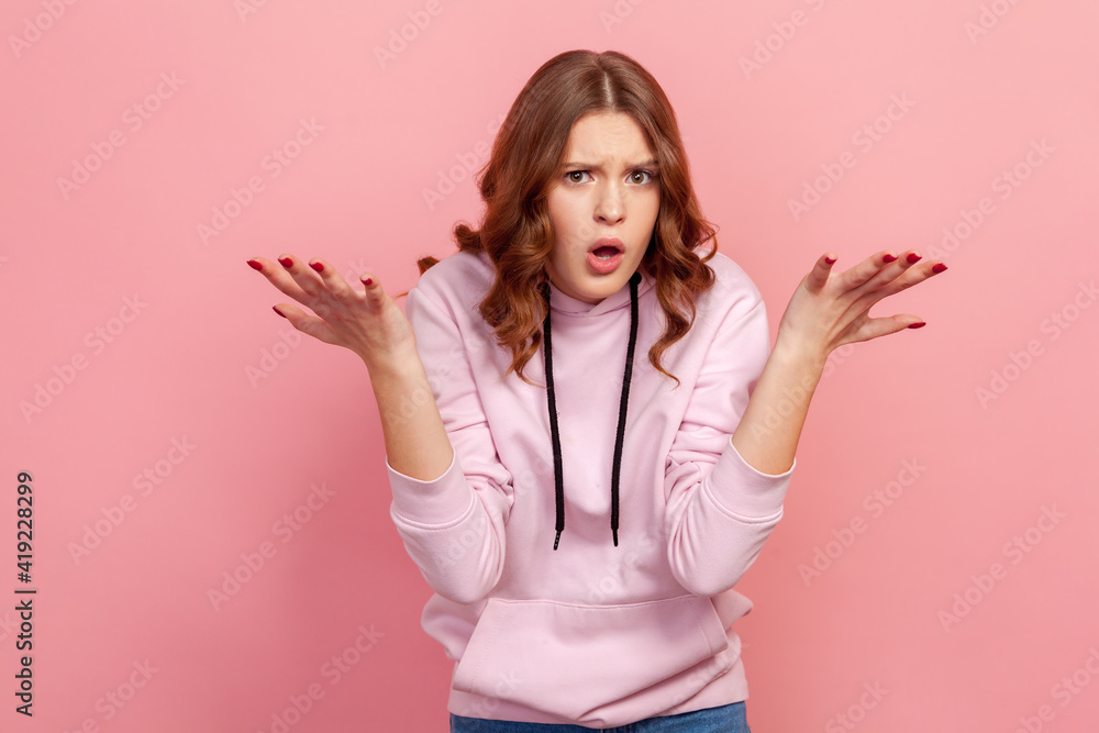 Fototapeta premium Portrait of frustrated young brunette female in hoodie spreading hands with shock and misunderstanding, what do you want gesture. Indoor studio shot isolated on pink background