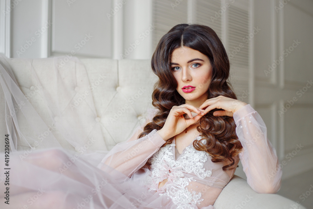 A bride in a white interior in a beautiful boudoir dress. She is sitting on the sofa, she has a beautiful wedding hairstyle and makeup A bride in a white interior in a beautiful boudoir dress. She is sitting on the sofa, she has a beautiful wedding hairstyle and makeup