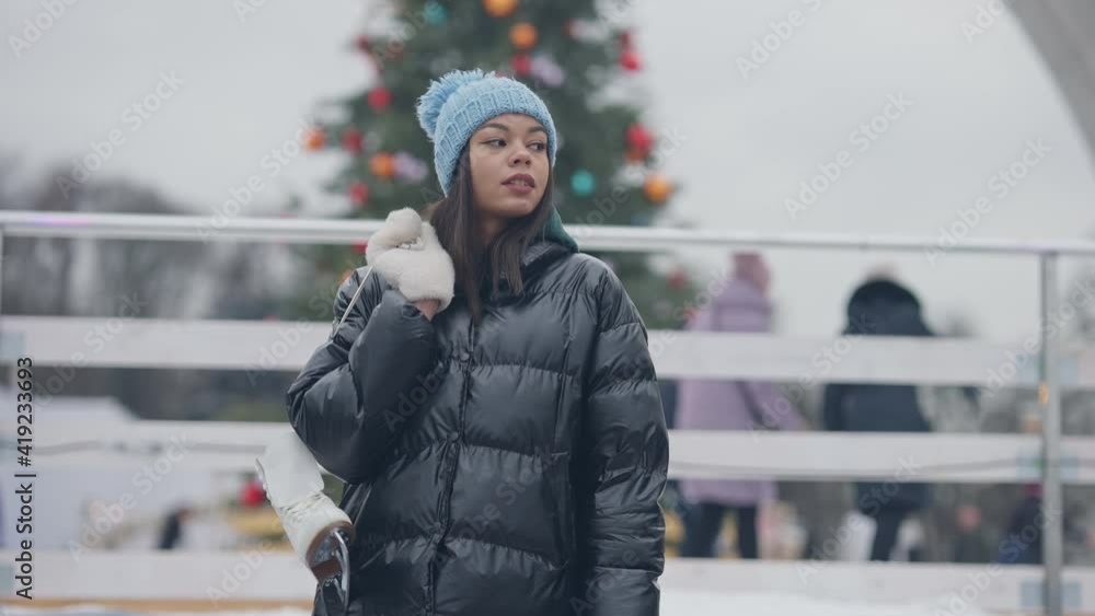 Portrait of confident millennial hanging ice skates on shoulder looking around on snowy winter day. Young beautiful African American woman resting outdoors with blurred ice rink at background. Portrait of confident millennial hanging ice skates on shoulder looking around on snowy winter day. Young beautiful African American woman resting outdoors with blurred ice rink at background.