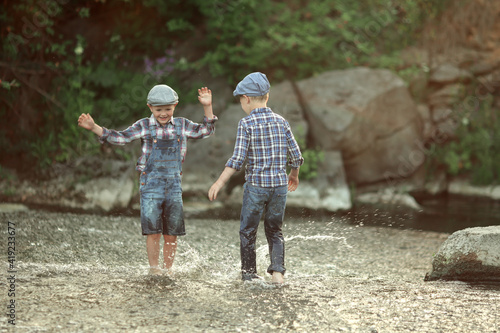 two little boys in denim overalls and a plaid shirt and caps having fun in the water and sprinkles