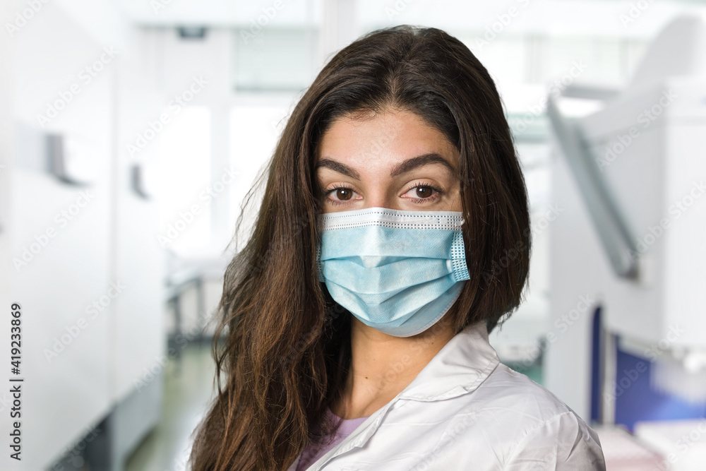 A brunette scientific researcher in a surgical gown and mask. In the background the blurred medical laboratory.