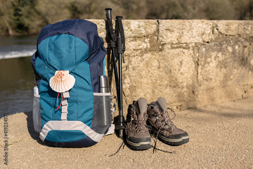 Canvastavla Backpack with seashell symbol of Camino de Santiago, trekking boots and poles leaning on stone wall