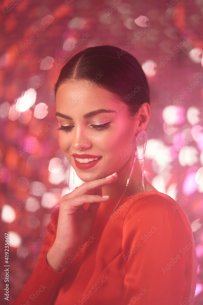 Beautiful lady in red sexy dress with make-up and elegant hairstyle. Pretty brunette woman posing against shining sequins background. Party concept. Colorful pink light Beautiful lady in red sexy dress with make-up and elegant hairstyle. Pretty brunette woman posing against shining sequins background. Party concept. Colorful pink light