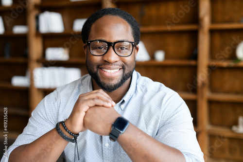 Headshot of friendly and confident African-American guy wearing stylish eyeglasses and smart casual wear, looks at the camera and holds hands in the lock near chin. Employee profile photo