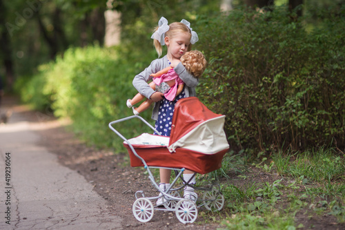 Cute smiling girl with bows in a blue dress playing with her red toy stroller and doll in the park . Childhood in the Soviet Union