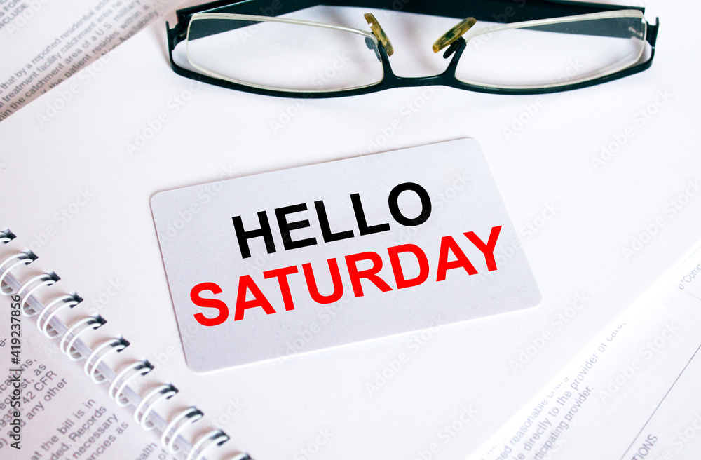 Text Hello Saturday on a business card lying on a notepad with eyeglasses and text documents Text Hello Saturday on a business card lying on a notepad with eyeglasses and text documents