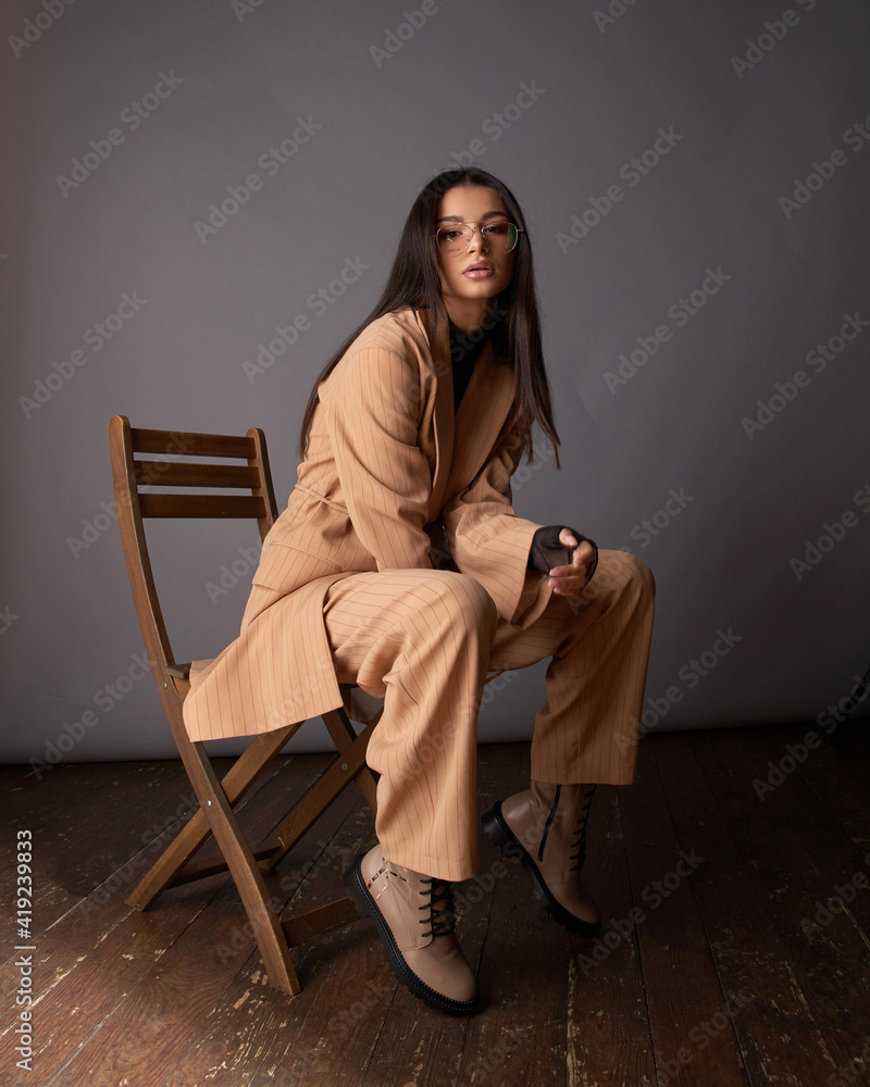 Young elegant woman in beige classical costume sitting at wooden chair against grey background. Pretty female model with natural make-up and long straight black hair Young elegant woman in beige classical costume sitting at wooden chair against grey background. Pretty female model with natural make-up and long straight black hair