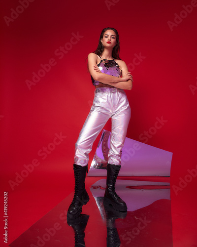Fashionable stylish female model wearing silver jeans, sequins top and black boots with perfect makeup and wet brunette hair standing and posing against red background