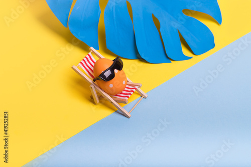 Creative funny composition with Easter egg with sunglasses while sitting on deck chair on illuminating yellow and pastel blue background