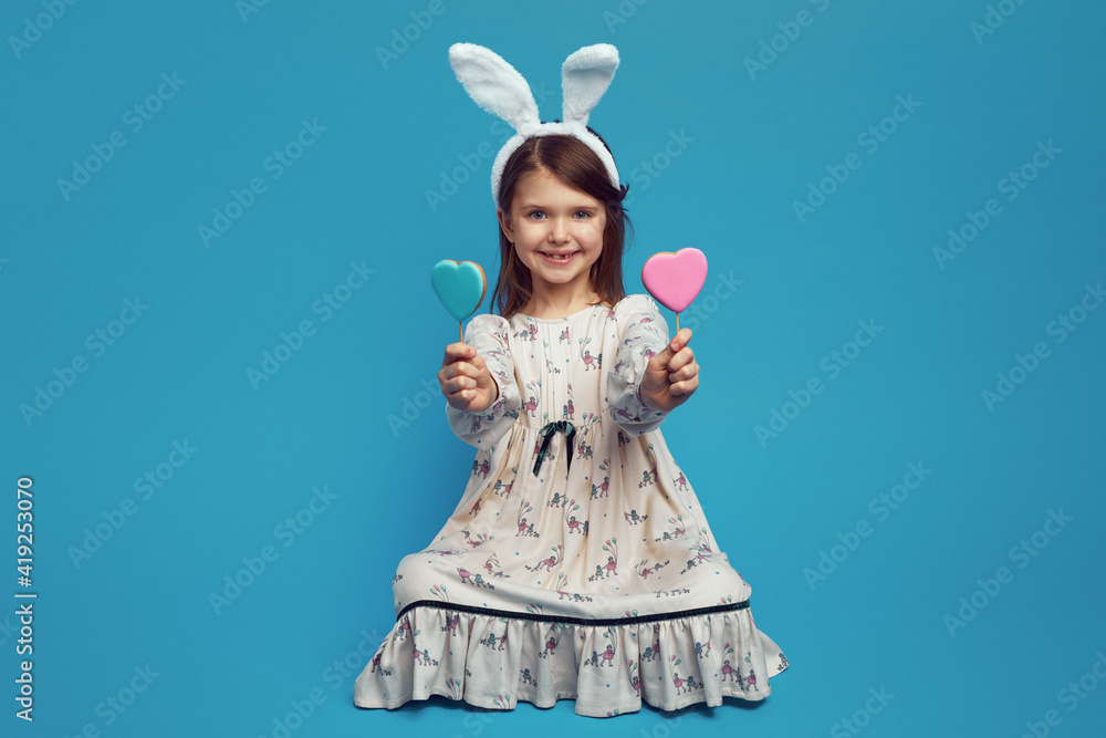 Nice cute kid girl having two cookies in heart shape on sticks, wearing easter bunny ears, isolated over blue background