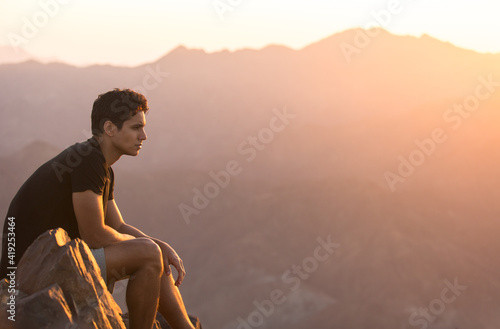 Vászonkép Attractive young man sitting alone relaxing and enjoying the view on top of a mountain peak at beautiful sunrise