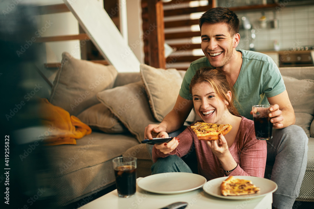 Happy couple watching something funny on TV while eating pizza at home.