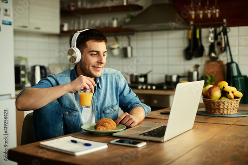 Smiling man with headphones surfing the net on laptop while working at home.