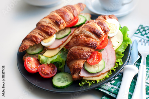 Croissant sandwich with ham, cheese, fresh salad, tomatoes, eggs and cucumber on black plate on white background. Breakfast with fresh croissant sandwich
