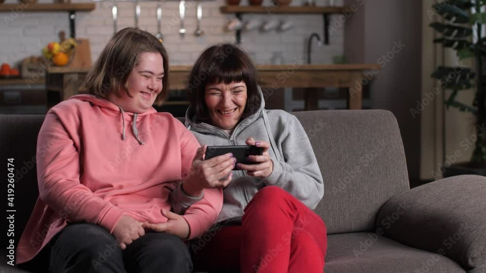 Close-up of cheerful disabled girl with down syndrome and her mom laughing while watching video on cellphone. Positive child with mother enjoying funny content on smartphone relaxing at home Close-up of cheerful disabled girl with down syndrome and her mom laughing while watching video on cellphone. Positive child with mother enjoying funny content on smartphone relaxing at home