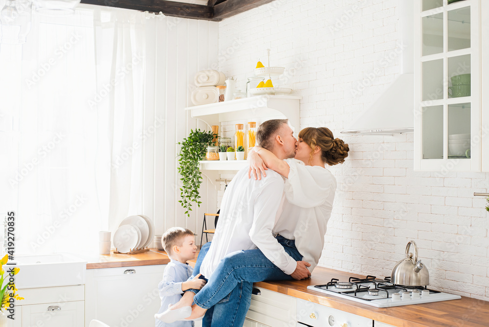 Young parents hug and kiss against the background of modern kitchen, a little son looks at them and hugs dad