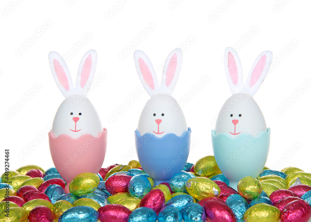 Row of Easter Eggs crafted into bunnies with felt ears, sitting in colorful egg cups with colorful foil wrapped chocolate egg candies, isolated on white background. Row of Easter Eggs crafted into bunnies with felt ears, sitting in colorful egg cups with colorful foil wrapped chocolate egg candies, isolated on white background.