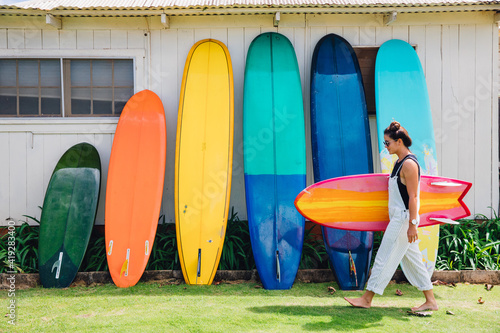 girl walking with surfboard in front of wall of longboards