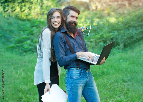 Business couple of woman and man with devices of laptop glasses mobile phone paper folder and standing outdoor on green grass.