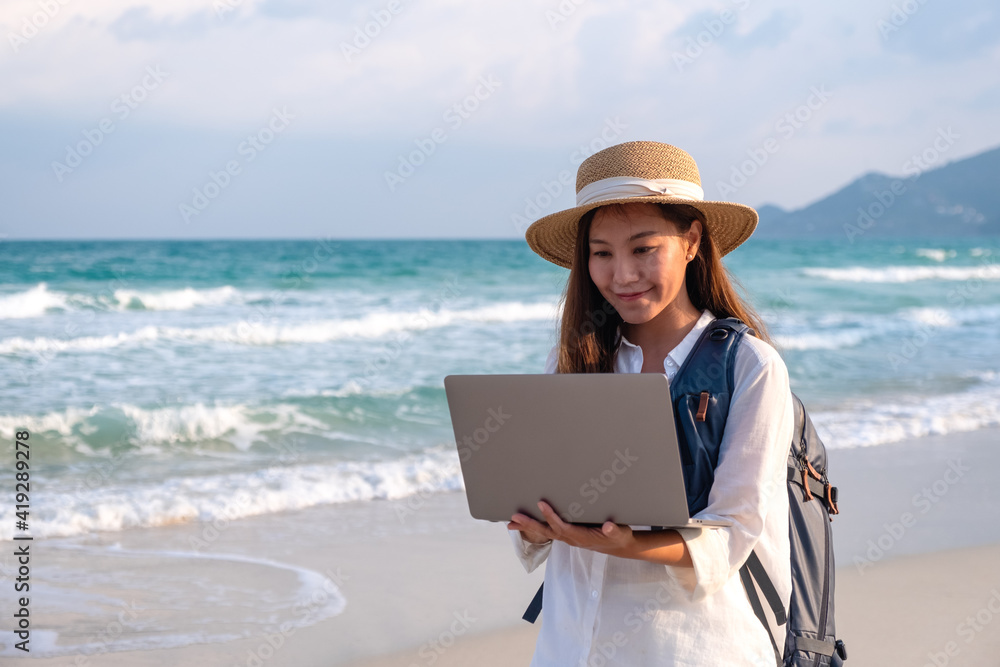 A young female traveler using and working on laptop computer while walking on the beach