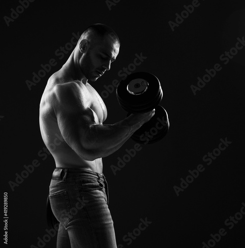 Muscular strong men, bodybuilder is working out, lifting dumbbells, doing exercises for biceps looking at it in gym over dark back ground with copy space. Young man lifting weights. Black and white 
