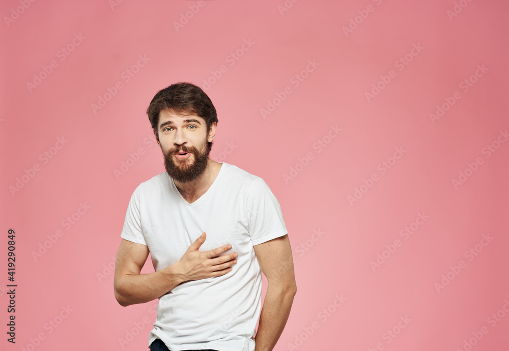 A man with a beard in a white T-shirt on a pink background gestures with his hands A man with a beard in a white T-shirt on a pink background gestures with his hands