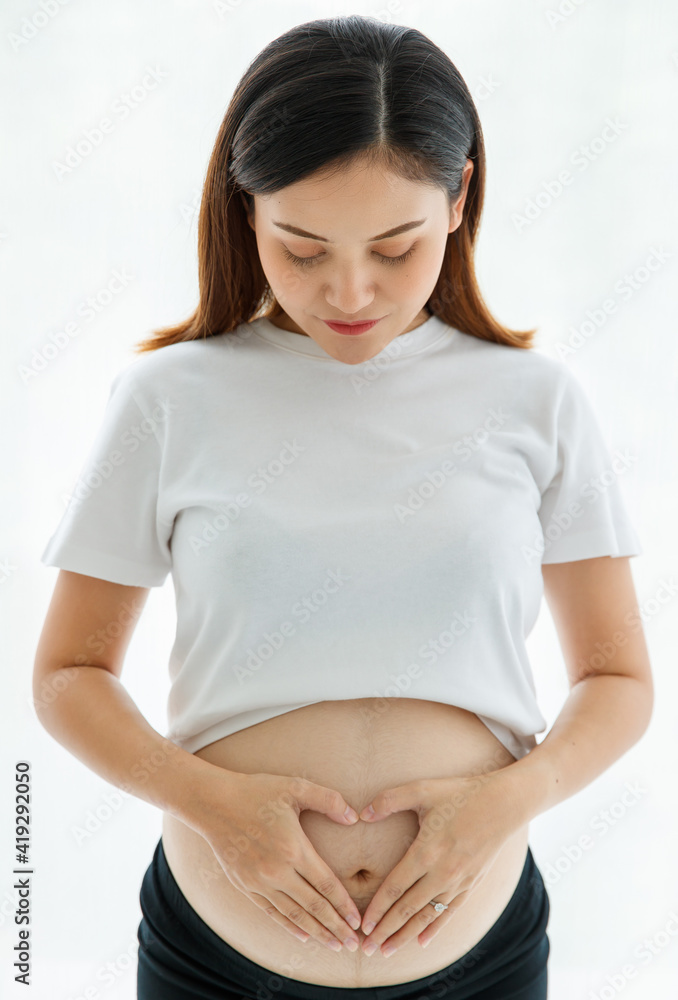 portrait of a young beautiful Asian pregnant woman with black hair wearing a casual dress standing and making a gesture of heart shape on her belly. Healthy and confident mother concept portrait of a young beautiful Asian pregnant woman with black hair wearing a casual dress standing and making a gesture of heart shape on her belly. Healthy and confident mother concept